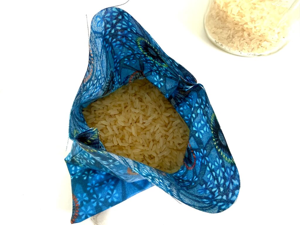 Open cloth bag with uncooked rice, blue patterned fabric, and a glass jar containing rice in the background on a white surface.