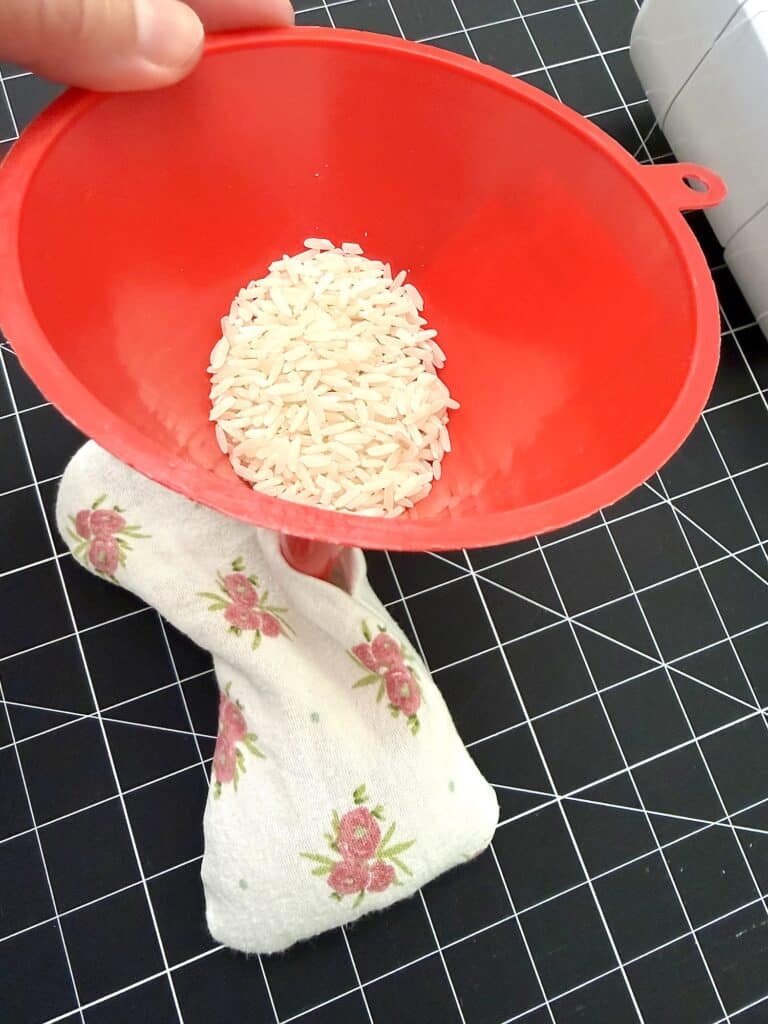 A hand holds a red funnel filled with uncooked rice, positioned above a small floral-patterned fabric pouch on a black grid cutting mat.