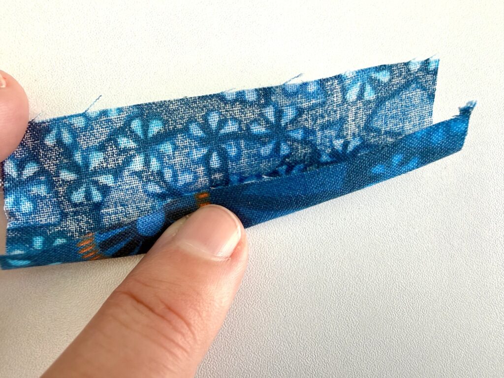 A hand holds a piece of blue patterned fabric, folding back one edge to show the reverse side against a white background.
