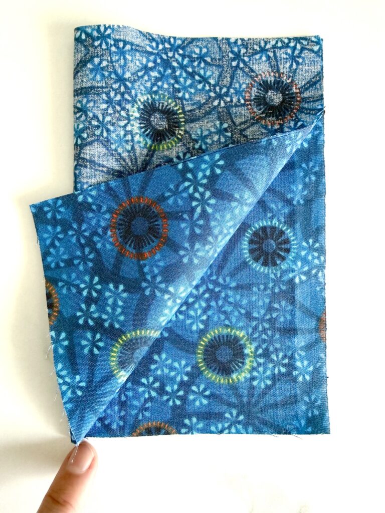 A hand lifts the corner of a folded piece of blue fabric with circular and starburst patterns on a white background.