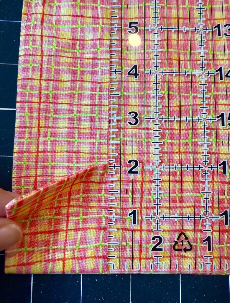 A hand folds a piece of yellow, pink, and orange plaid fabric next to a transparent quilting ruler marked with measurements, all on a black grid cutting mat.