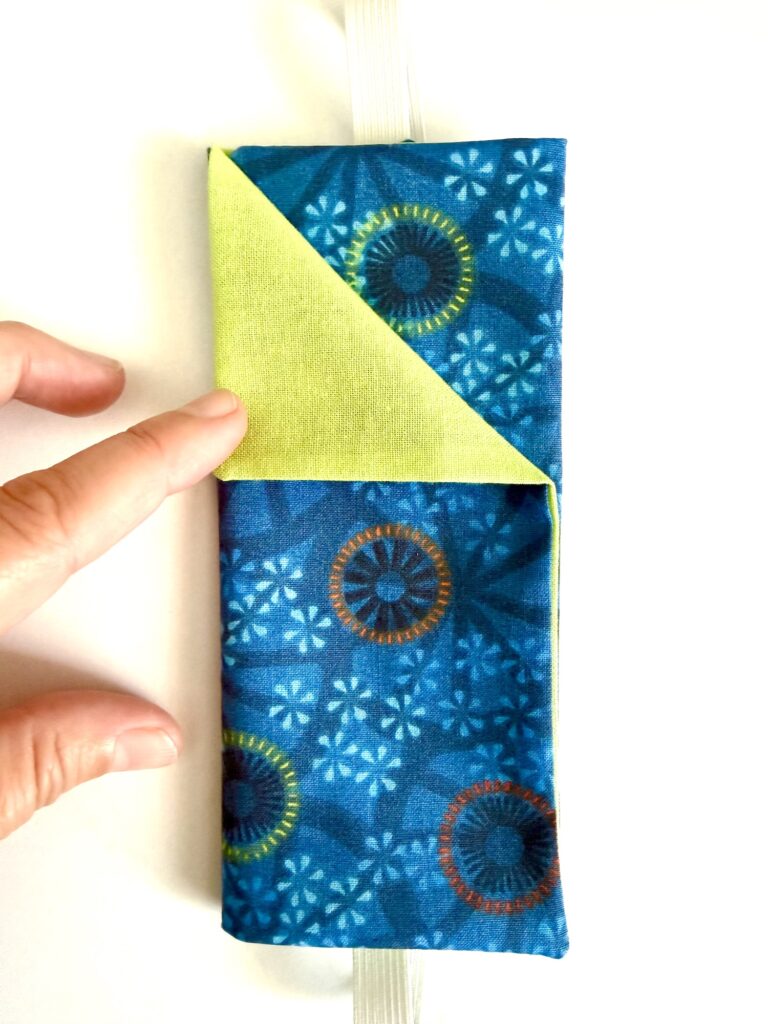 A hand folds the corner of a rectangular, blue and green patterned fabric item with a yellow inner lining against a white background.