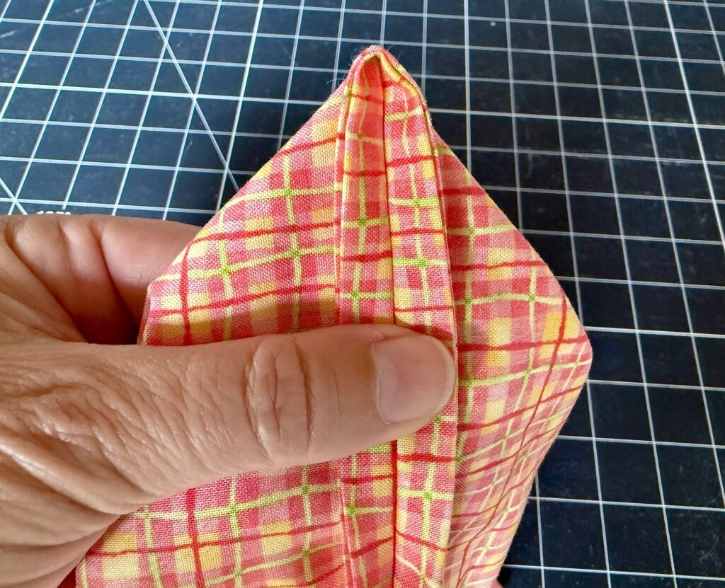 A hand holds a piece of colorful plaid fabric folded into a pointed shape, with a black cutting mat featuring a white grid in the background.