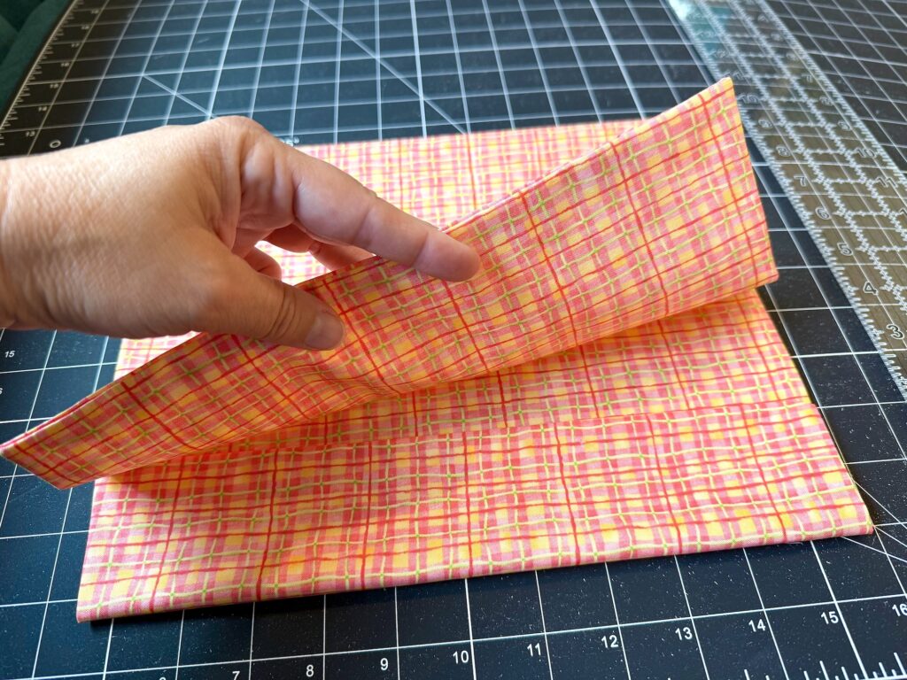 A hand lifts a folded piece of pink and yellow plaid fabric on a black grid cutting mat with a clear ruler nearby.