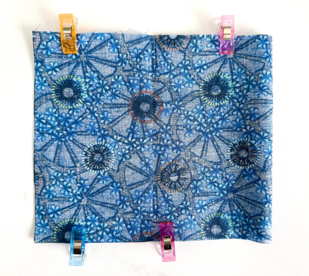 Square piece of blue patterned fabric held at four corners with colorful clips on a white background.