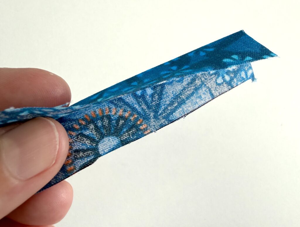 A hand holds a strip of blue patterned fabric, folded lengthwise with the raw edges aligned against a white background.