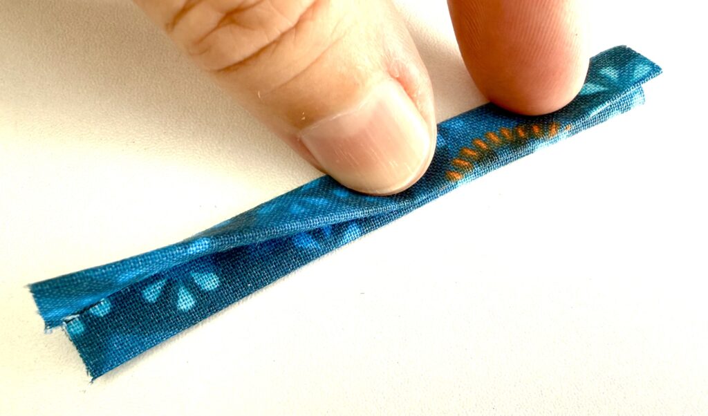 A hand holds a folded strip of blue patterned fabric with orange stitching, placed on a white surface.