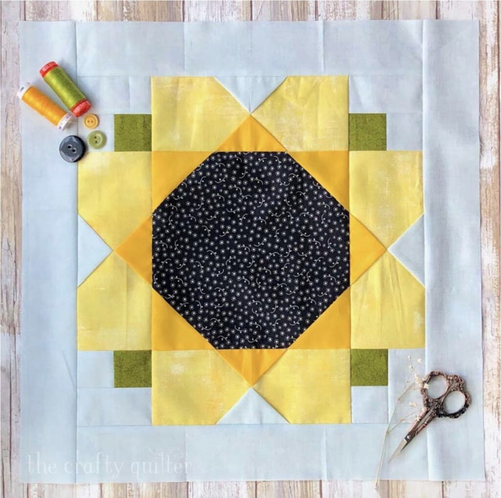 A sunflower quilt block with yellow petals, green corners, a black polka-dotted center, sewing thread, buttons, and scissors arranged on a light wooden surface.