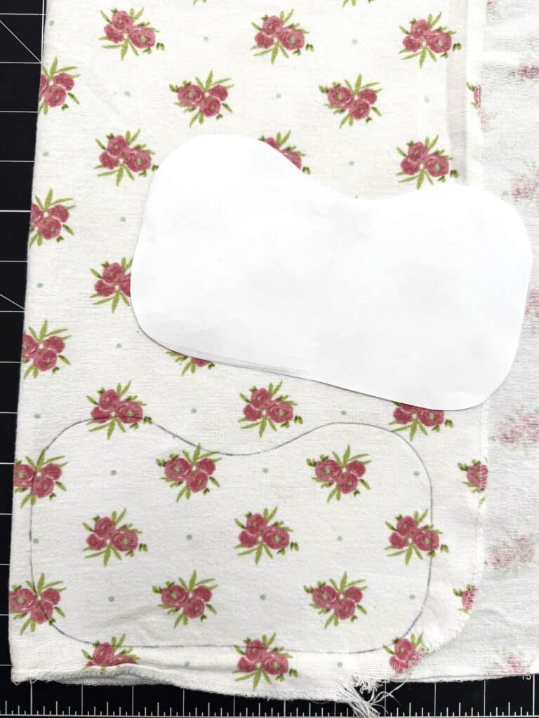 A piece of floral fabric with a pattern template placed on top and an outline traced onto the fabric, shown on a cutting mat.