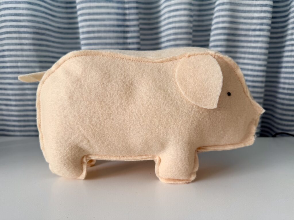 A beige plush pig toy stands on a white surface in front of blue and white striped curtains.