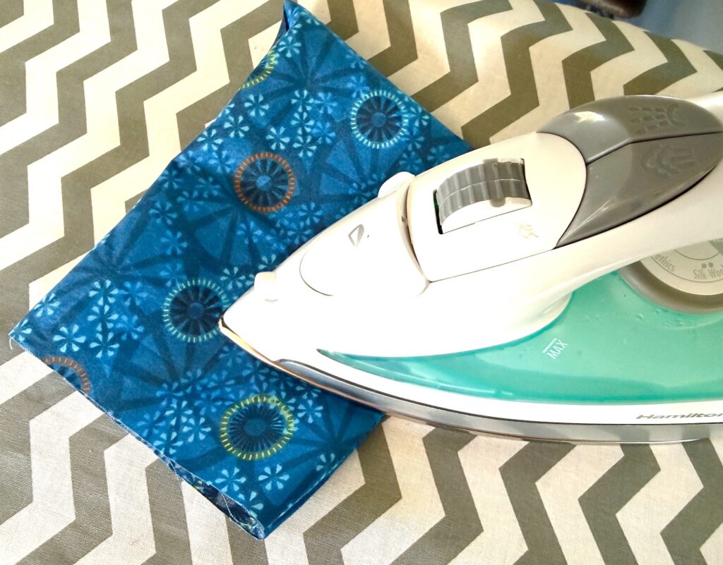 A white and teal clothes iron rests on a small, folded blue patterned fabric atop a chevron-patterned ironing board cover.