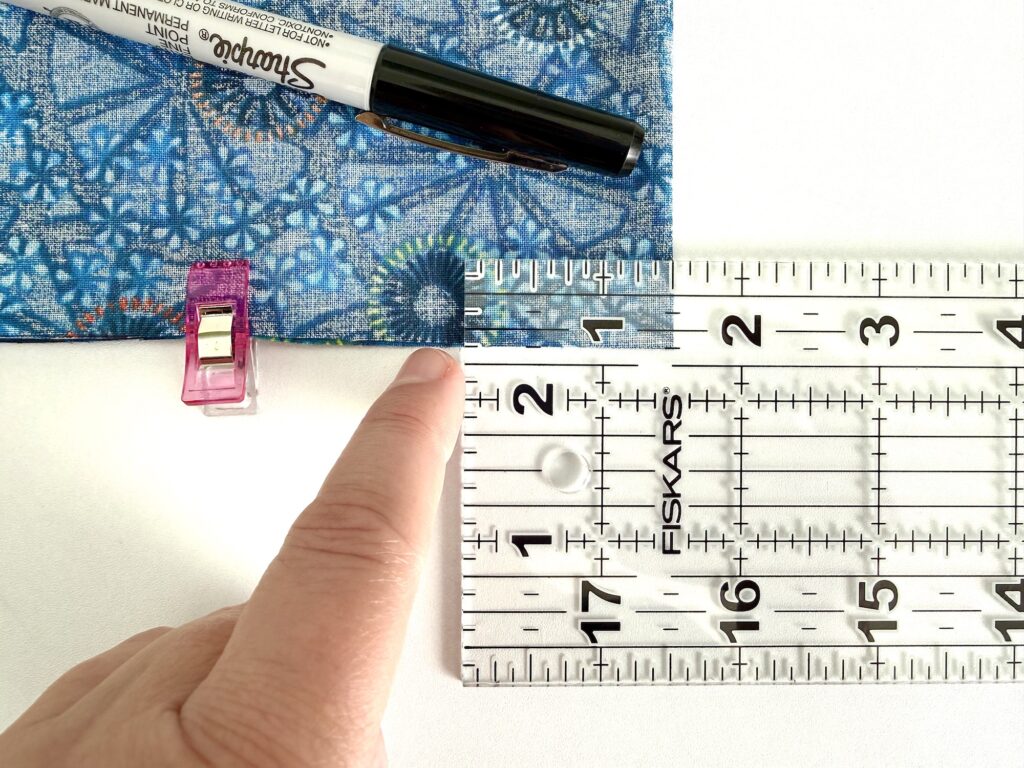 A hand holds a clear ruler against blue patterned fabric, marking a measurement with a black marker. A pink fabric clip is placed nearby.