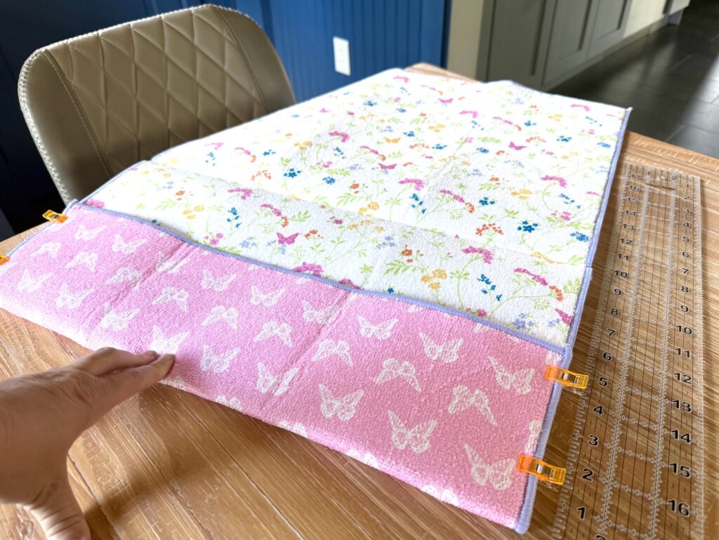 A hand lifts the corner of two stacked pieces of patterned fabric clipped together on a wooden table next to a clear ruler.