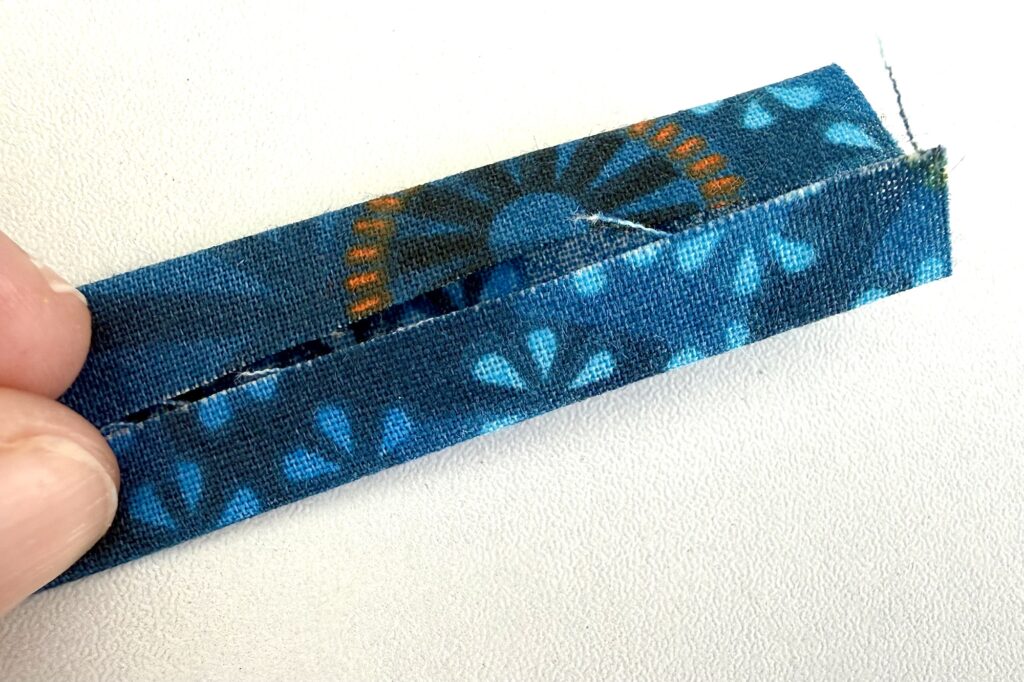 A hand holds a folded strip of blue patterned fabric with an uneven stitched seam along one edge, placed on a white surface.