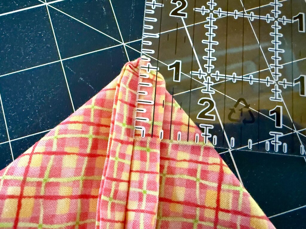 A piece of folded plaid fabric is positioned under a clear acrylic ruler on a black cutting mat with a white grid.