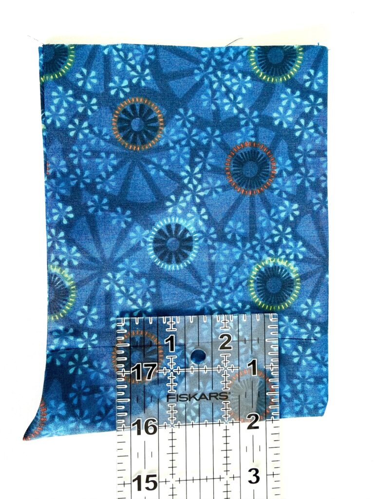 A piece of blue patterned fabric with circular and geometric designs is measured with a clear ruler, showing a width of about 3.5 inches.