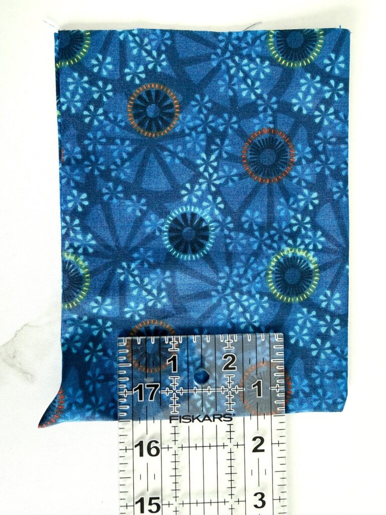 A blue patterned fabric square is measured with a ruler showing about 4 inches by 5 inches. The fabric has circular and starburst designs in various blue shades.