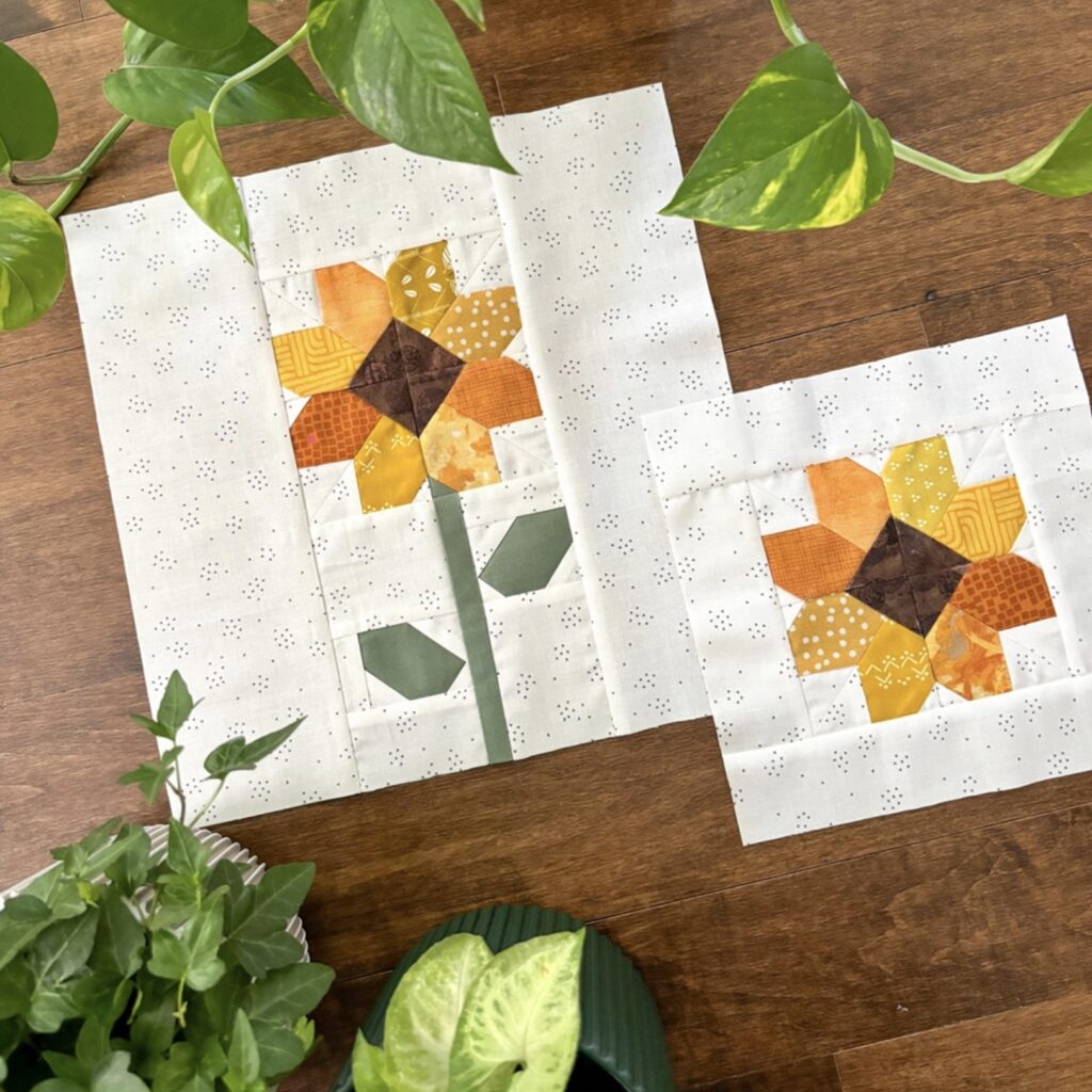 Two quilt blocks with yellow and orange patchwork flower designs are displayed on a wooden surface, surrounded by green potted plants.