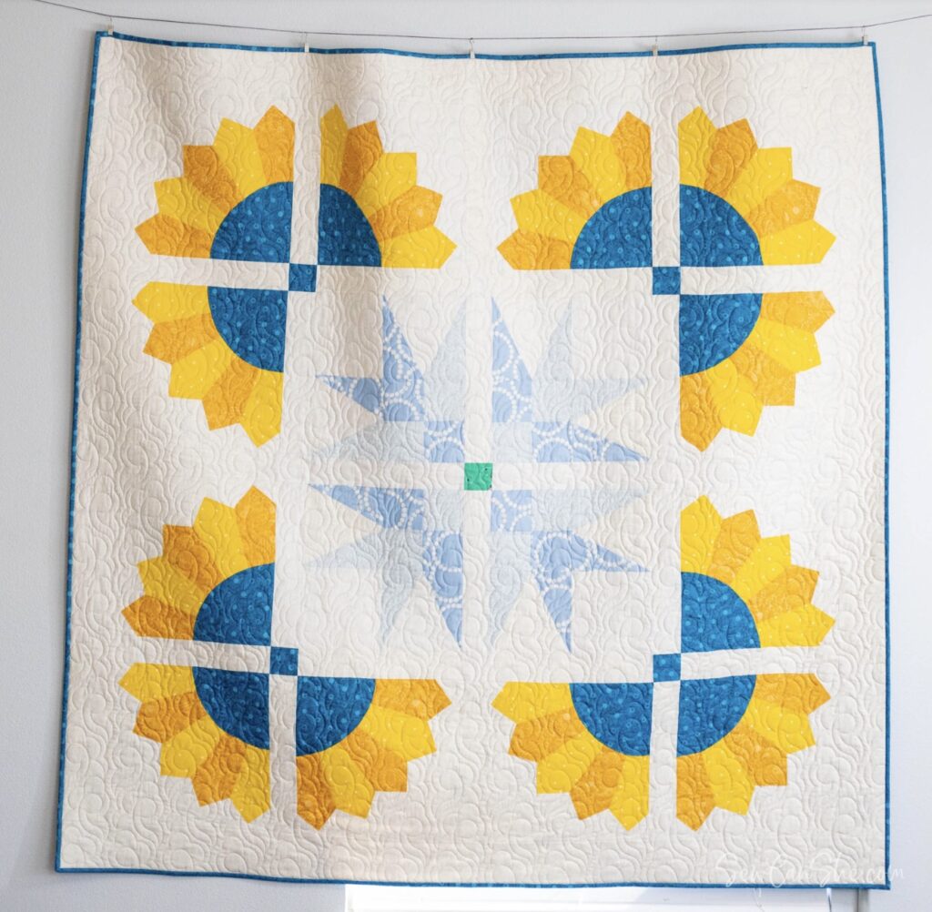 A quilt featuring four yellow and blue sunflower patterns in each corner and a light blue star design in the center on a white background.