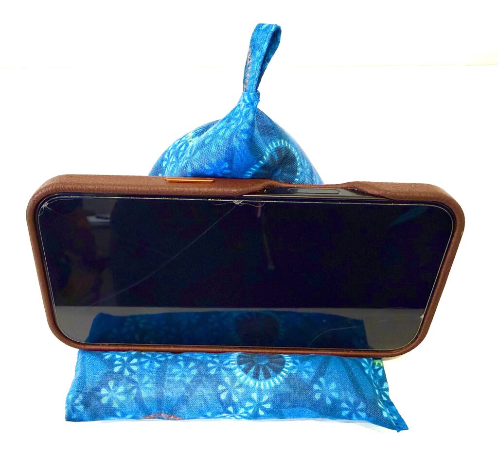 A smartphone in a brown case rests horizontally on a blue fabric beanbag stand with a loop, against a white background.