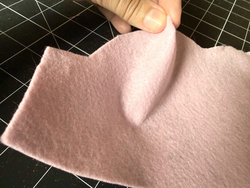 A hand lifts the edge of a piece of light pink felt, showing its texture. The felt is placed on a black grid cutting mat.