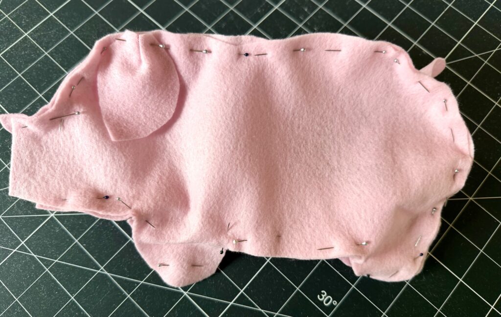 Two pieces of pink felt cut in the shape of a pig are pinned together with sewing pins, placed on a black gridded cutting mat.
