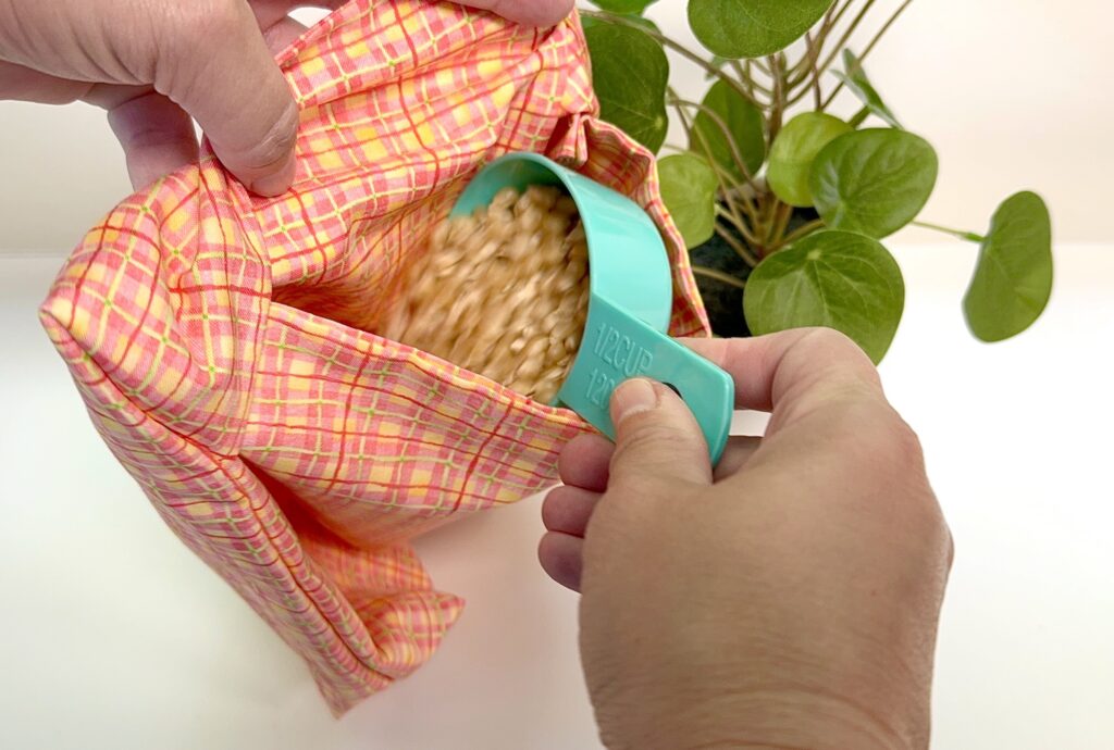 A hand pours uncooked popcorn kernels from a blue measuring cup into a pink plaid fabric bag, with a green plant in the background.