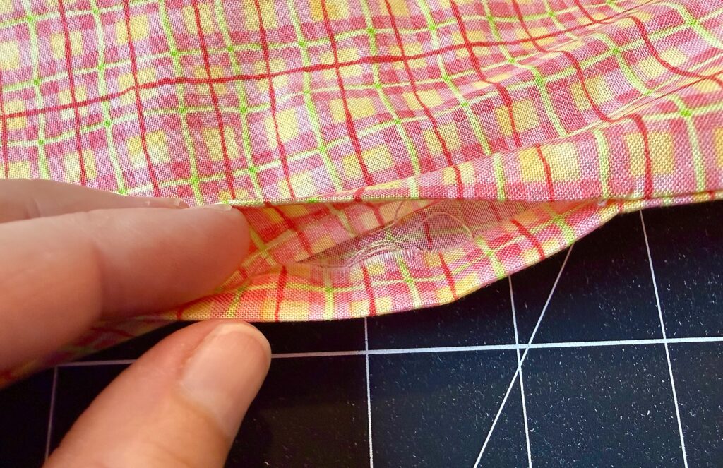A hand holds the edge of a pink, yellow, and orange plaid fabric, revealing a small, discreetly sewn pocket against a black grid background.