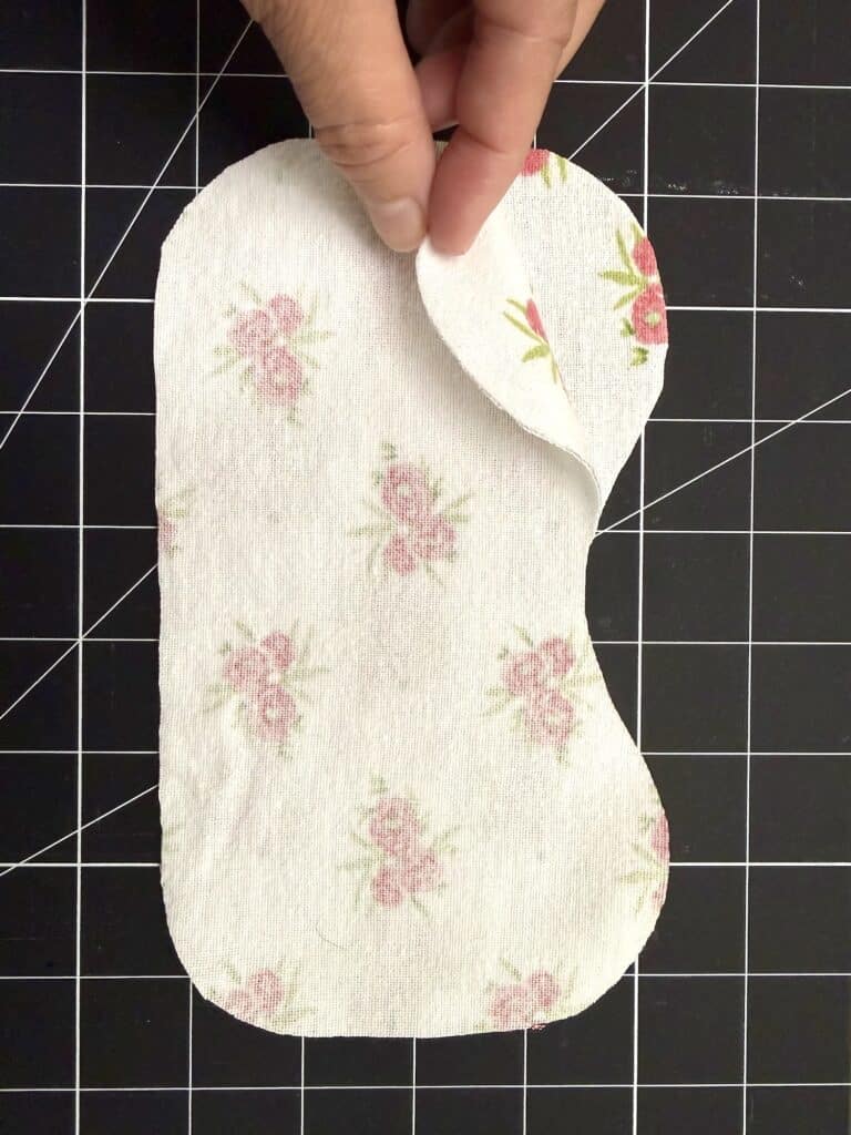 A hand lifts the top layer of a fabric pad with a floral pattern, placed on a black grid cutting mat.