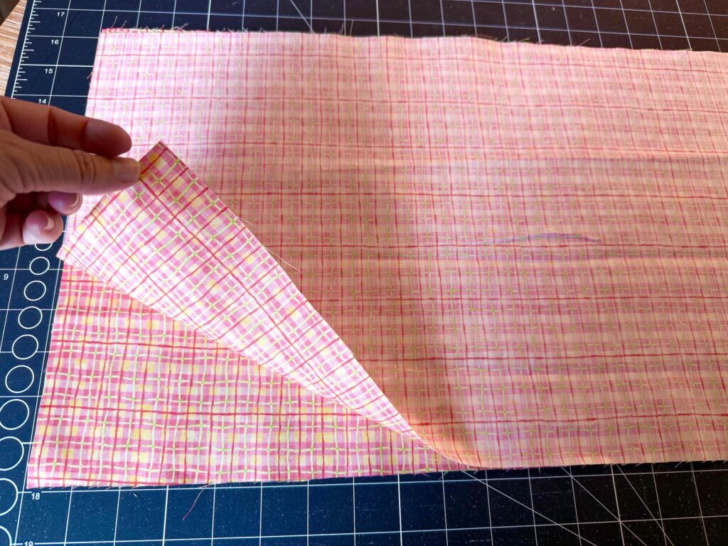 A hand lifts the corner of pink plaid fabric on a cutting mat with measurement lines visible in the background.