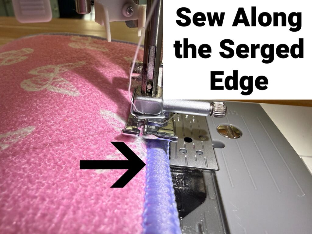 A sewing machine stitching along the serged edge of pink fabric, with text instructing to sew along the serged edge and a black arrow pointing to the seam.