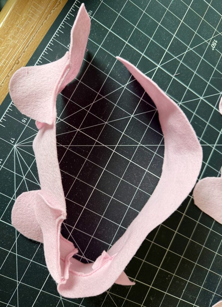 A pink felt fabric piece with curved and pointed edges is laid on a black cutting mat with a white grid pattern.