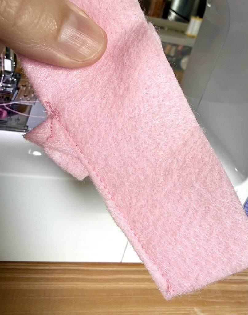 A hand holds a piece of pink felt fabric with a straight machine-sewn seam, next to a sewing machine on a wooden surface.