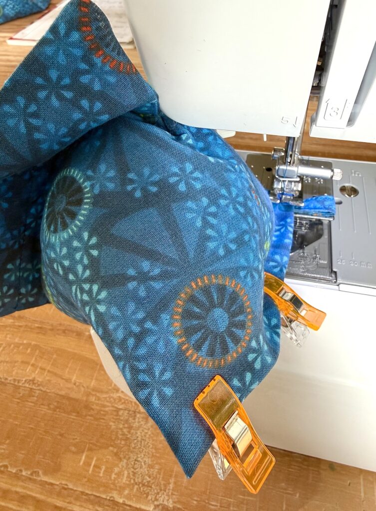 Blue patterned fabric, partially sewn on a sewing machine, is held together with orange plastic clips on a wooden surface.