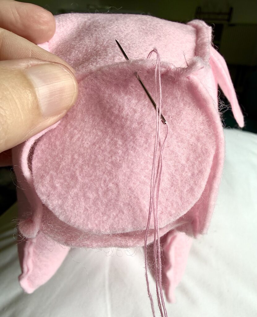 A hand holds pink felt pieces while a needle with pink thread is used to sew a circular piece onto a stuffed object.