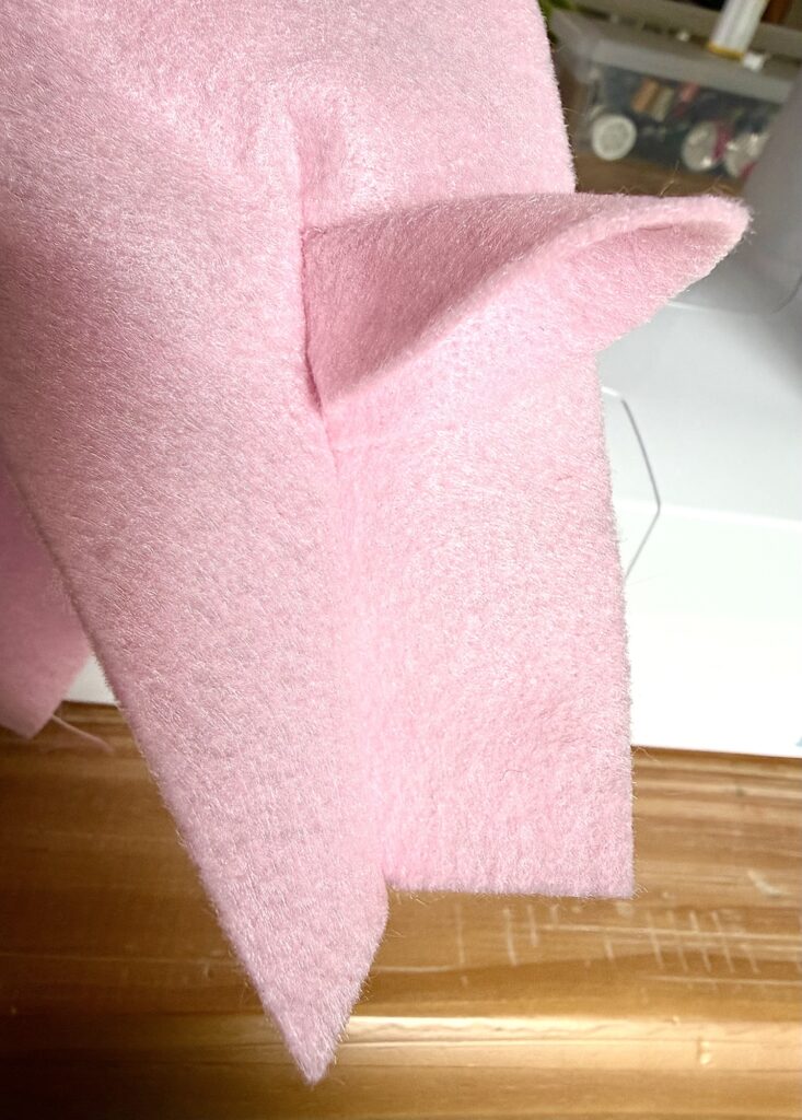 A close-up of a piece of light pink felt fabric with sewn-in triangular ear shapes, positioned on a wooden surface near a sewing machine.