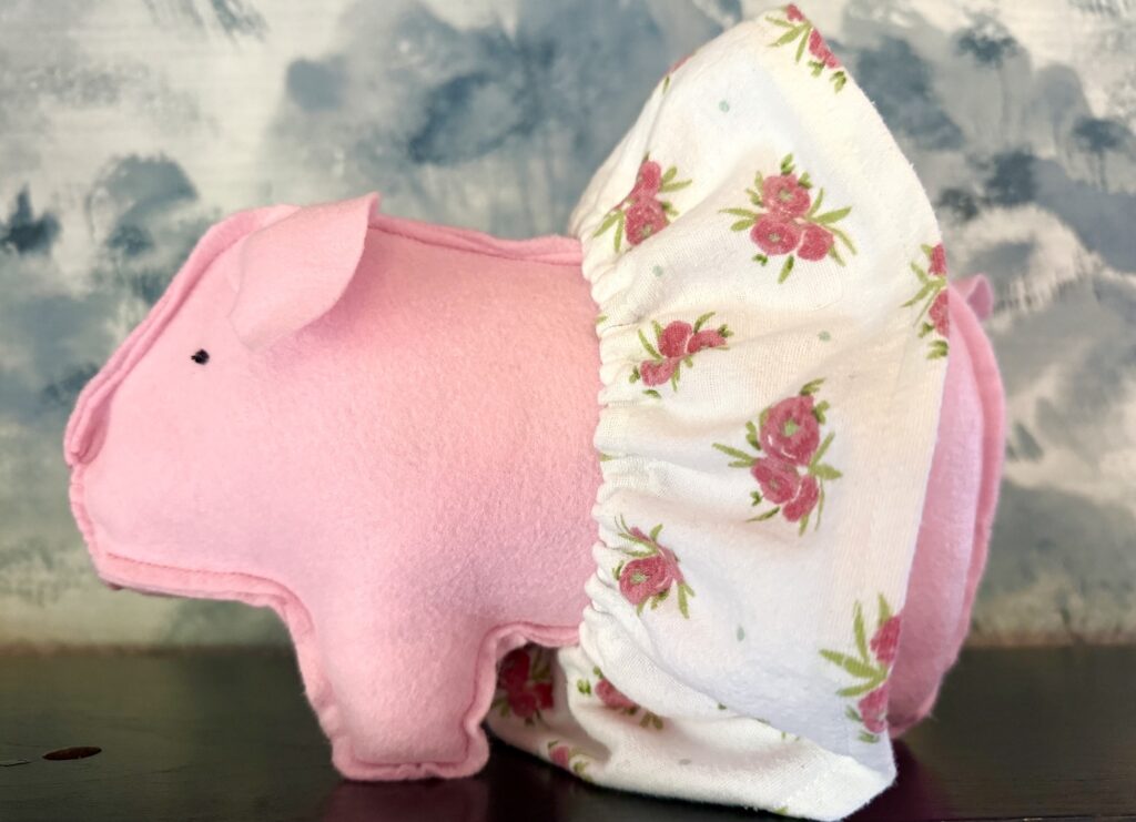 A pink plush pig with stitched features wears a white floral-patterned skirt. The background is an abstract mix of blue and white tones.