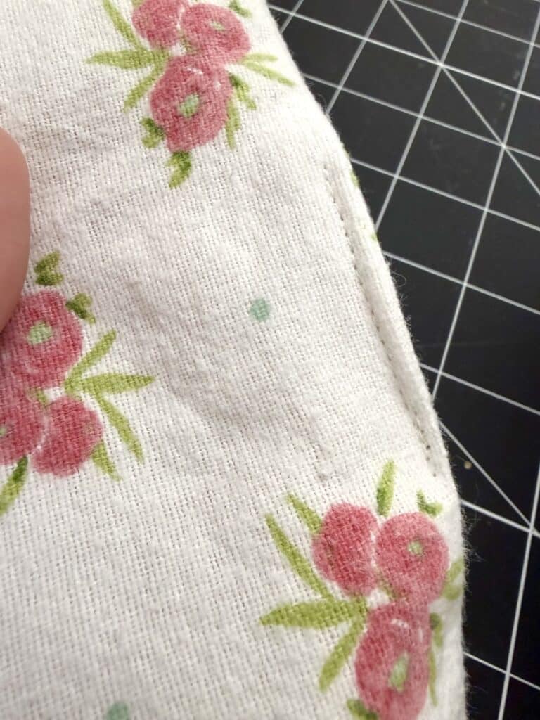 Close-up of white fabric with pink and green floral print, showing a stitched seam. A black cutting mat with a grid pattern is partially visible in the background.