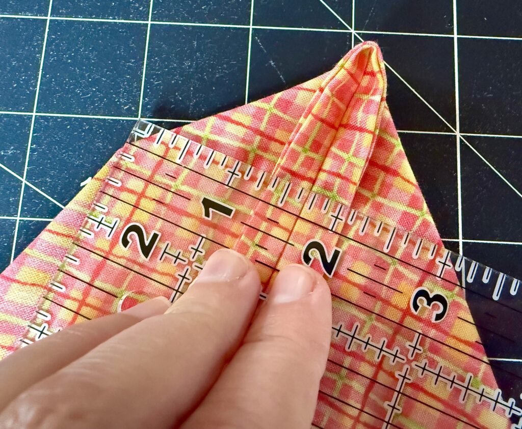 A hand holds a clear ruler against a folded piece of plaid fabric on a gridded cutting mat, measuring the seam.