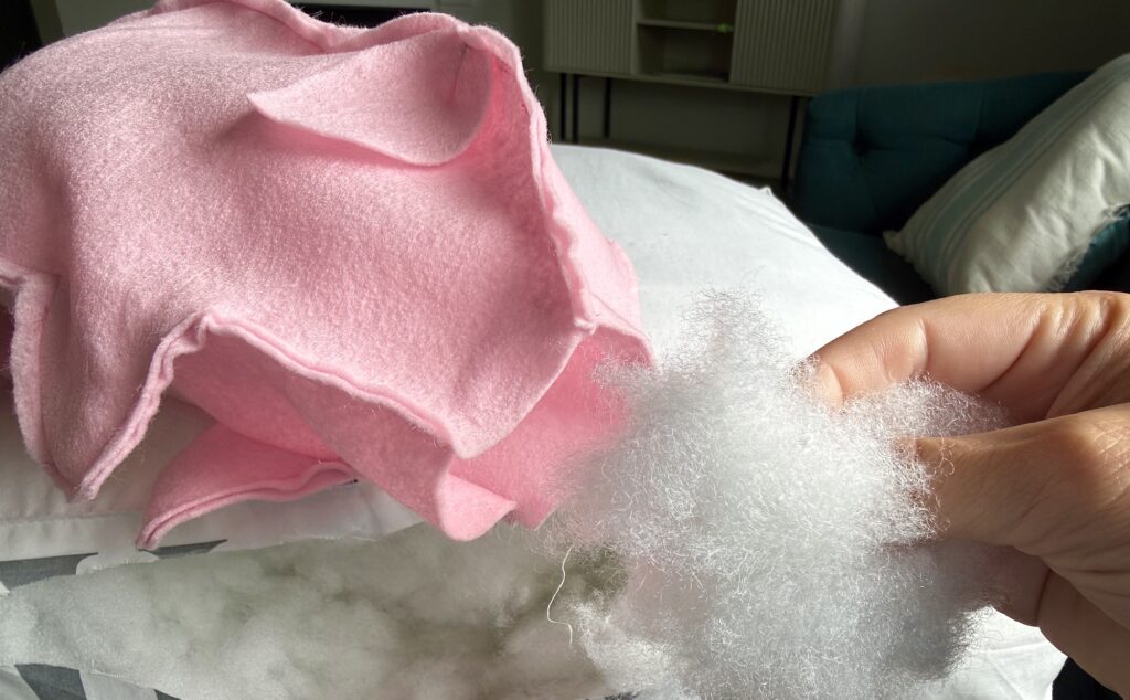 A hand holds white polyester stuffing near a pink fabric piece, possibly for a sewing or crafting project, with a pillow and couch in the background.