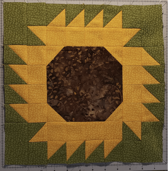A quilt block depicting a sunflower, with a brown center, yellow petals, and a green background, arranged in a geometric pattern on a gridded surface.