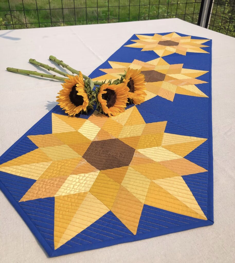 A quilted table runner with large sunflower patterns in yellow and brown on a blue background, displayed with three fresh sunflowers on a white table.