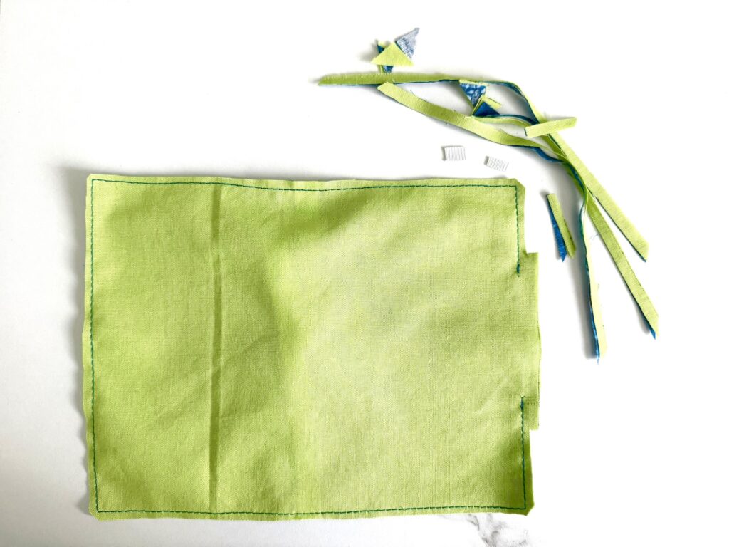 Rectangular piece of green fabric with sewn edges lies on a white surface; trimmed fabric strips and threads are nearby.