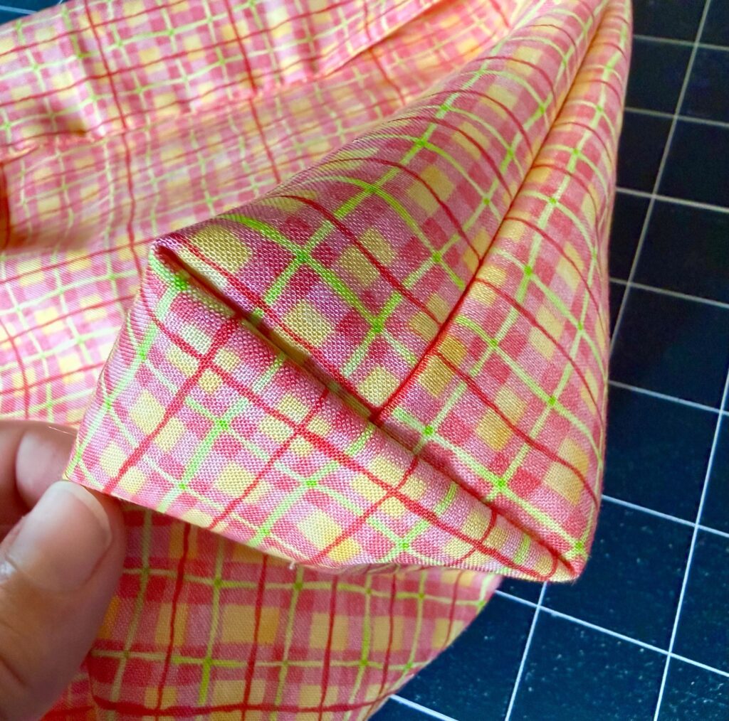 A hand holds a piece of fabric with a red, yellow, and green plaid pattern, folded precisely on a cutting mat.