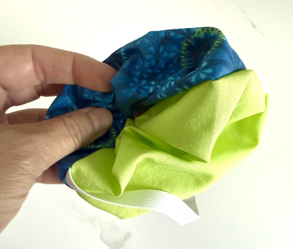 A hand holds a small fabric item with blue and green sections and a white elastic band, against a white background.