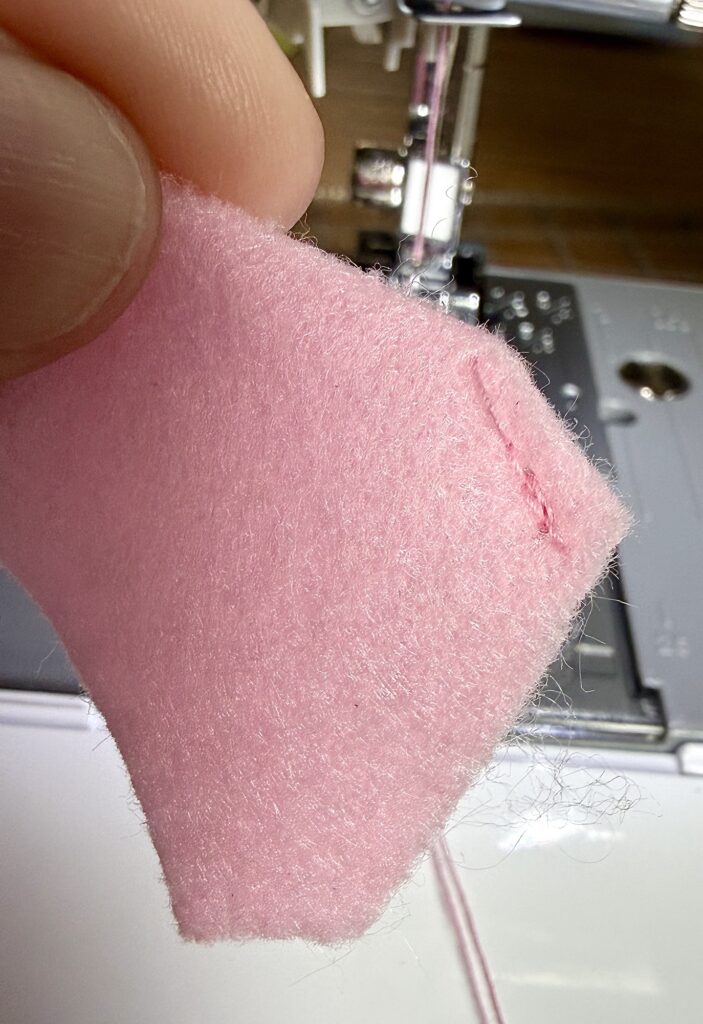 Close-up of a hand holding a piece of pink felt with a short line of pink stitching next to a sewing machine.