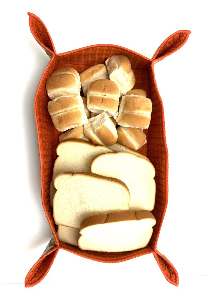an orange colored handmade drying mat bread basket filled with rolls and sandwich bread displayed against a white background