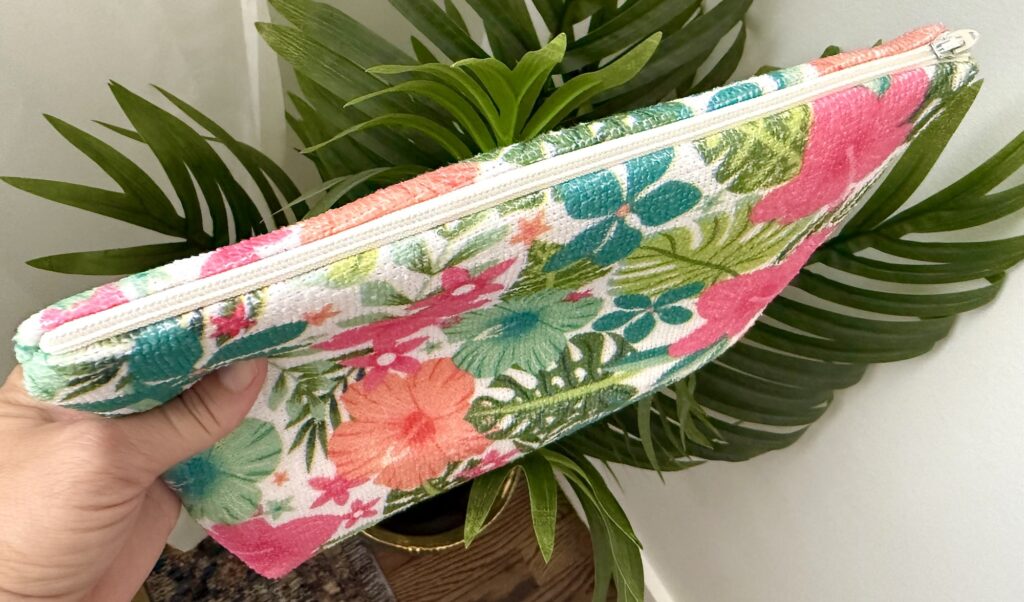 A hand holds a zippered pouch with a colorful floral pattern, positioned in front of large green leaves and a white wall.