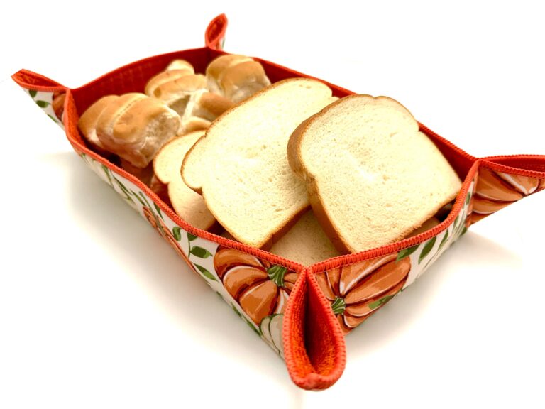 a handmade drying mat bread basket decorated with pumpkins is filled with rolls and sandwich bread displayed against a white background