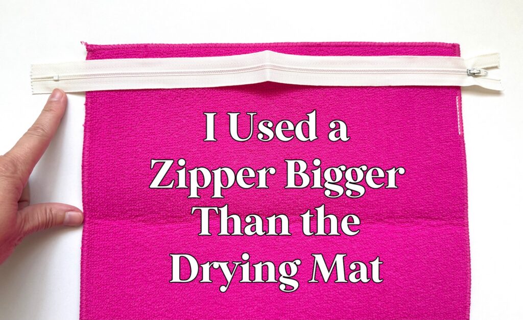 A hand points to a white zipper pinned to bright pink fabric with the text, “I Used a Zipper Bigger Than the Drying Mat.”.